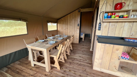 Interior of a safari tent with a wooden dining set, tableware, colorful mugs, and rustic wooden decor.