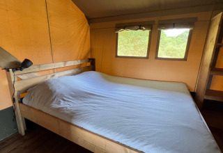 Interior view of a safari tent bedroom featuring a wooden-framed double bed and windows overlooking nature.