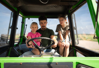 Ein Mann mit zwei Kindern sitzt in einem grünen Traktor bei FarmCamps Mariekerke, einem Ferienpark in Zeeland.