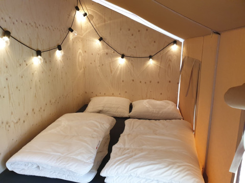 Vista interior de una tienda safari con dos camas individuales, ropa de cama blanca y guirnalda de luces.