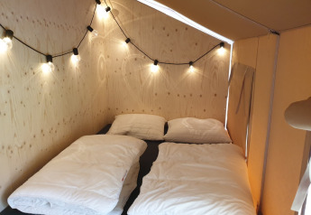 Innenaufnahme eines Safarizelts mit zwei Einzelbetten, weißer Bettwäsche und Lichterkette an der Wand.