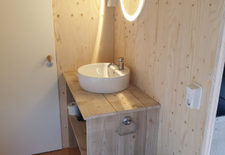 Bagno con pareti in legno, lavandino rotondo, specchio illuminato e bidone in una tenda safari nei Paesi Bassi.