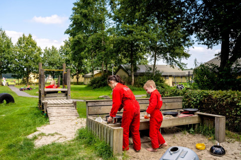 Børn leger i sandkasse på FarmCamps t Looveld i Drenthe, Nederlandene, omgivet af grøn natur og telte.