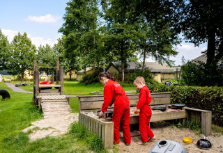 Børn leger i sandkasse på FarmCamps t Looveld i Drenthe, Nederlandene, omgivet af grøn natur og telte.