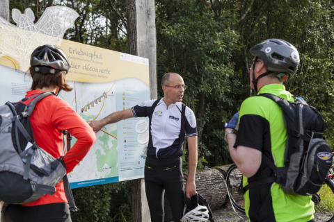 Three cyclists examine a trail map at Camping Troisvierges, Luxembourg, surrounded by lush green trees.