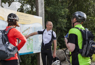 Three cyclists examine a trail map at Camping Troisvierges, Luxembourg, surrounded by lush green trees.