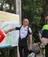 Tres ciclistas consultan un mapa en Camping Troisvierges, Luxemburgo, rodeados de vegetación y árboles.
