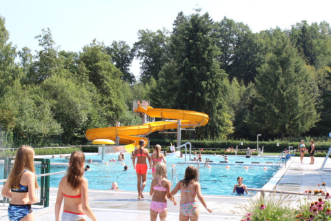 Niños y adultos disfrutan de la piscina al aire libre y tobogán amarillo en Camping Troisvierges, Luxemburgo.