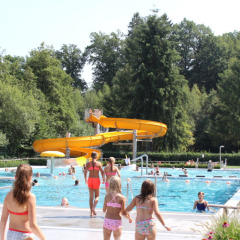 Niños y adultos disfrutan de la piscina al aire libre y tobogán amarillo en Camping Troisvierges, Luxemburgo.