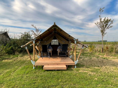 Safari tent called Duolodge with wooden porch, two chairs in front, and grass field under blue sky.