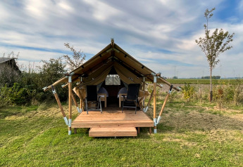 Safari tent called Duolodge with wooden porch, two chairs in front, and grass field under blue sky.