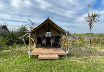 Safari-Zelt namens Duolodge mit Holzveranda, zwei Stühlen davor und grüner Wiese unter blauem Himmel.
