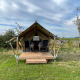 Safari telt kaldet Duolodge med træveranda, to stole foran og grøn eng i baggrunden under en blå himmel.