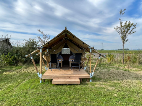 Tienda safari llamada Duolodge con porche de madera, dos sillas al frente y campo verde bajo cielo azul.