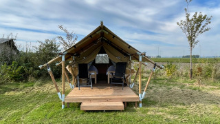 Safari tent called Duolodge with wooden porch, two chairs in front, and grass field under blue sky.