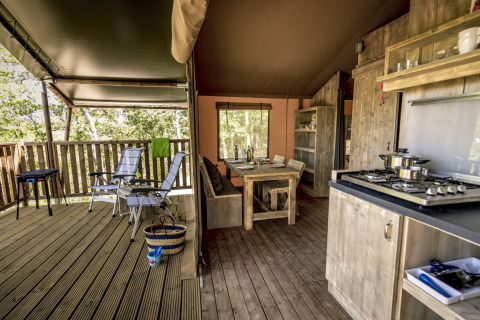 Interior of a safari tent featuring wooden furniture, open terrace with chairs, and a full kitchen area.