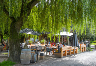 Udendørs caféområde under store træer på Camping Mareveld, Limburg, Holland, med gæster der nyder solen.