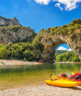 Colorful kayaks on a riverbank with a natural stone arch and mountains near Gluiras, Auvergne-Rhône-Alpes, France.