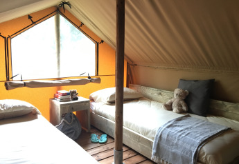 Interno di una tenda di glamping accogliente con due letti, finestra grande, comodino e orsacchiotto.