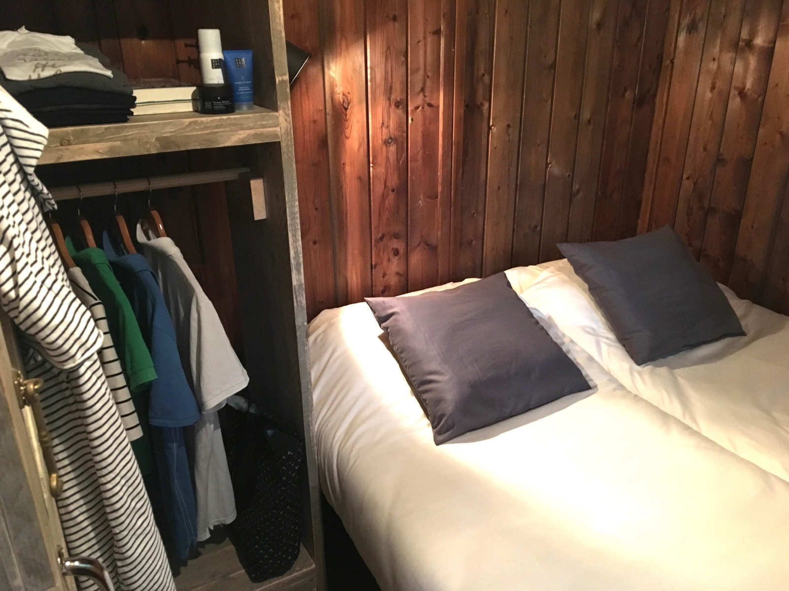 Knusse glampingkamer met houten wanden, wit opgemaakt tweepersoonsbed en een open kledingkast.