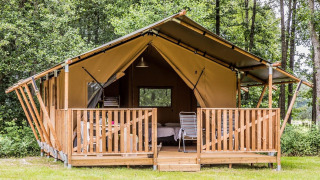 Tenda safari con veranda in legno in mezzo al bosco, sedie da esterno e zona letto visibile all'interno.