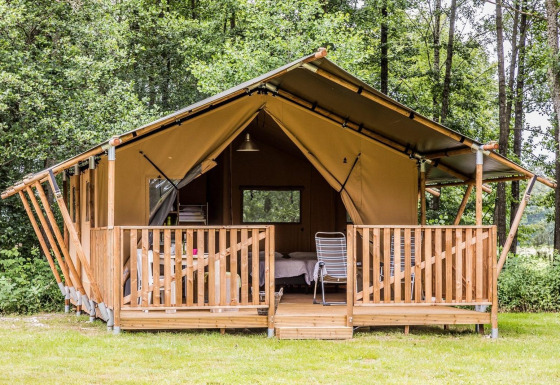 Tienda safari con terraza de madera en un entorno de bosque, con sillas y cama visible en el interior.