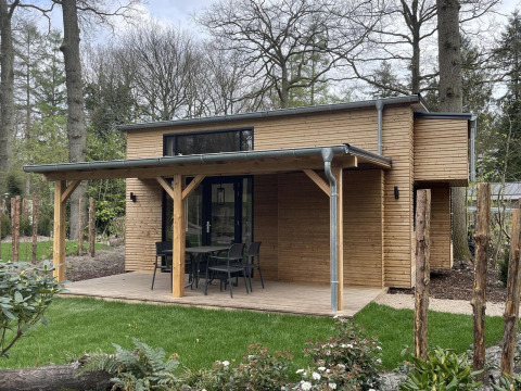 Pequeña casa moderna de madera con terraza cubierta, muebles de exterior y jardín verde alrededor.
