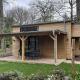 Et moderne tiny house i træ med overdækket terrasse og havemøbler, omgivet af grøn have og træer.