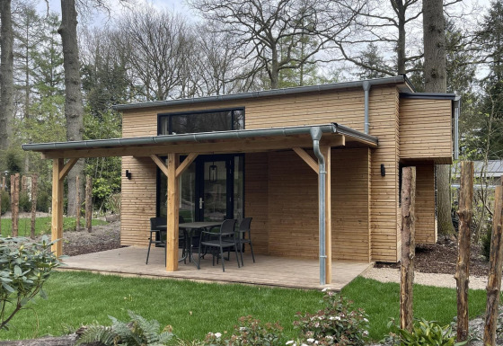 Klein modern huisje in hout met overdekt terras, tuinmeubilair en omringd door groen en bomen.
