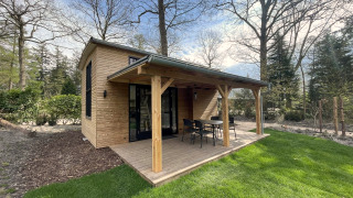 Lille træhus med overdækket terrasse og havemøbler omgivet af grøn græsplæne og træer om foråret.