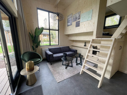Moderne tiny house woonkamer met houten muren, zwarte bank, groene stoel, raam en trap naar de slaapzolder.