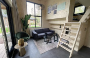 Modern tiny house living room with wood walls, loft ladder, black sofa, green chair, and large windows.