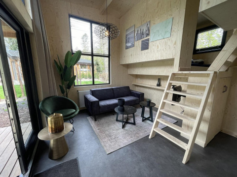 Moderne tiny house stue med sort sofa, grøn stol, trævægge, trappe og store vinduer mod haven.