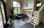 Moderne tiny house stue med sort sofa, grøn stol, trævægge, trappe og store vinduer mod haven.
