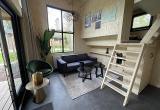 Moderne tiny house stue med sort sofa, grøn stol, trævægge, trappe og store vinduer mod haven.
