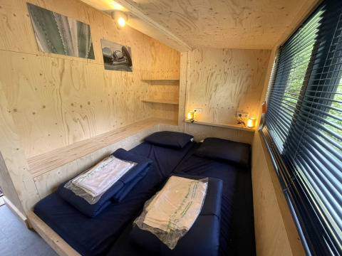 Tiny house slaapkamer met tweepersoonsbed, houten wanden, raam met jaloezieën en sfeerverlichting.