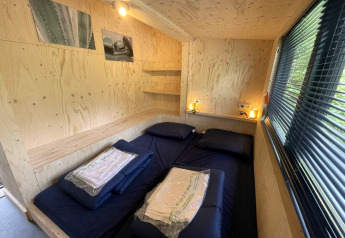 Dormitorio en casa pequeña con cama doble, paredes de madera, lámparas, ventana y ambiente acogedor.