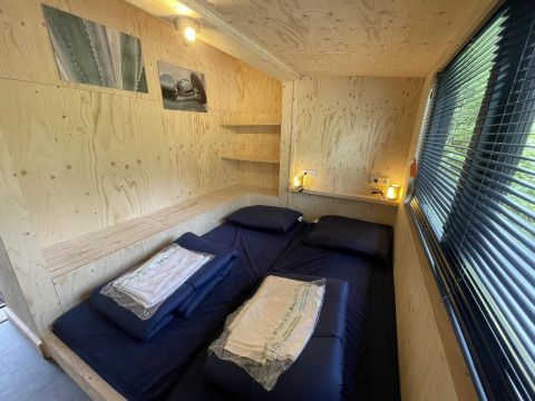 Chambre d’une tiny house avec lit double, murs en bois, stores à la fenêtre et éclairage chaleureux.
