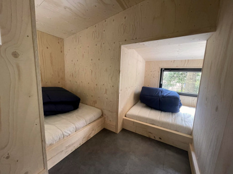 Twee bedden met blauwe dekens in een tiny house met lichte houten wanden en raam met boszicht.