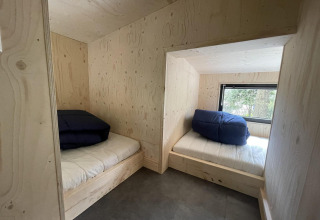 To senge med blå tæpper i et lille hus med lyse trævægge og et vindue, der vender ud mod skoven.