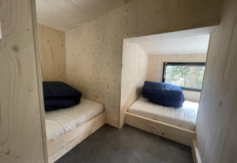 Due letti con coperte blu in una tiny house con pareti in legno chiaro e finestra sul bosco.