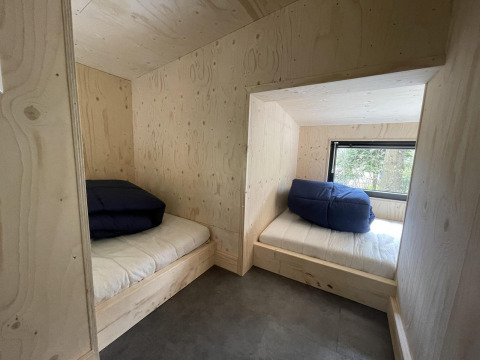 Due letti con coperte blu in una tiny house con pareti in legno chiaro e finestra sul bosco.