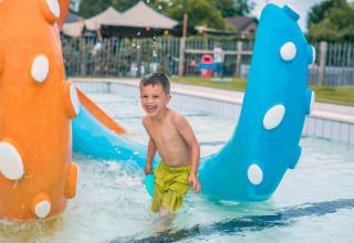 Niño sonriente jugando en una piscina al aire libre con juguetes de colores cerca de una tiny house.