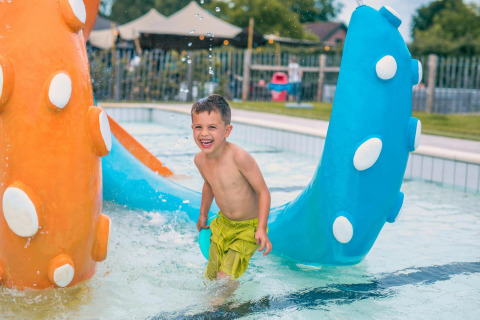 Bambino felice gioca in una piscina all'aperto vicino a una tiny house con giochi d'acqua colorati.