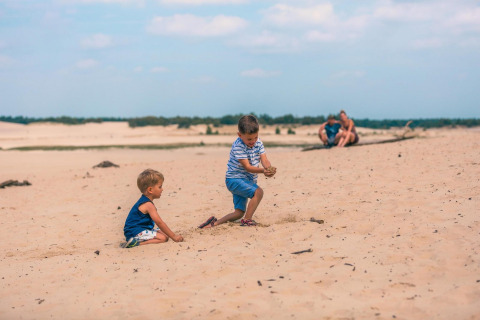 Zwei Kinder spielen im Sand bei Tiny House in Kaatsheuvel, Niederlande, Eltern sitzen im Hintergrund.