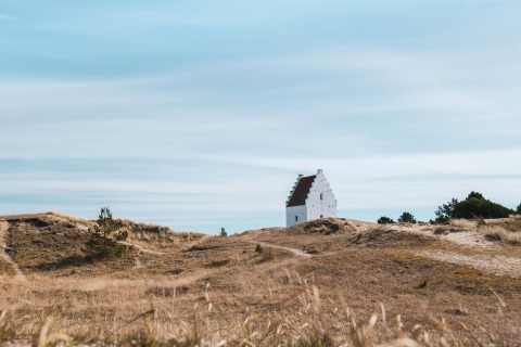 A solitary white building sits among grassy dunes under a blue sky near Hirtshals, North Jutland, Denmark.