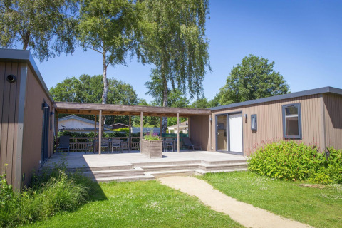 Photo extérieure de la Family Lodge avec terrasse en bois, mobilier de jardin et verdure sous un ciel ensoleillé.