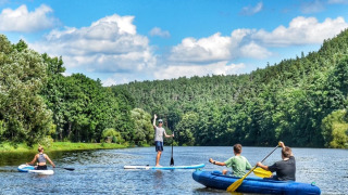 Folk padler på en flod omgivet af frodig skov ved Camping Prima i Sydbøhmen, Tjekkiet, under blå himmel.
