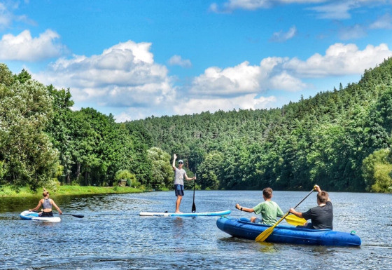 Folk padler på en flod omgivet af frodig skov ved Camping Prima i Sydbøhmen, Tjekkiet, under blå himmel.