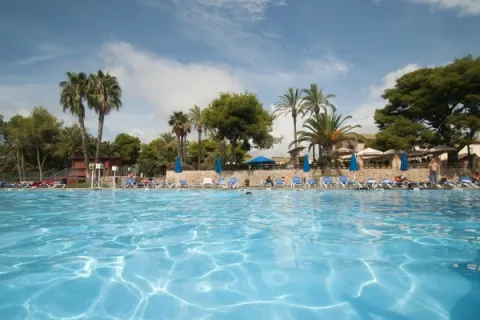 Large outdoor swimming pool with sun loungers, palm trees, and blue umbrellas at Vilanova Park, Catalonia, Spain.